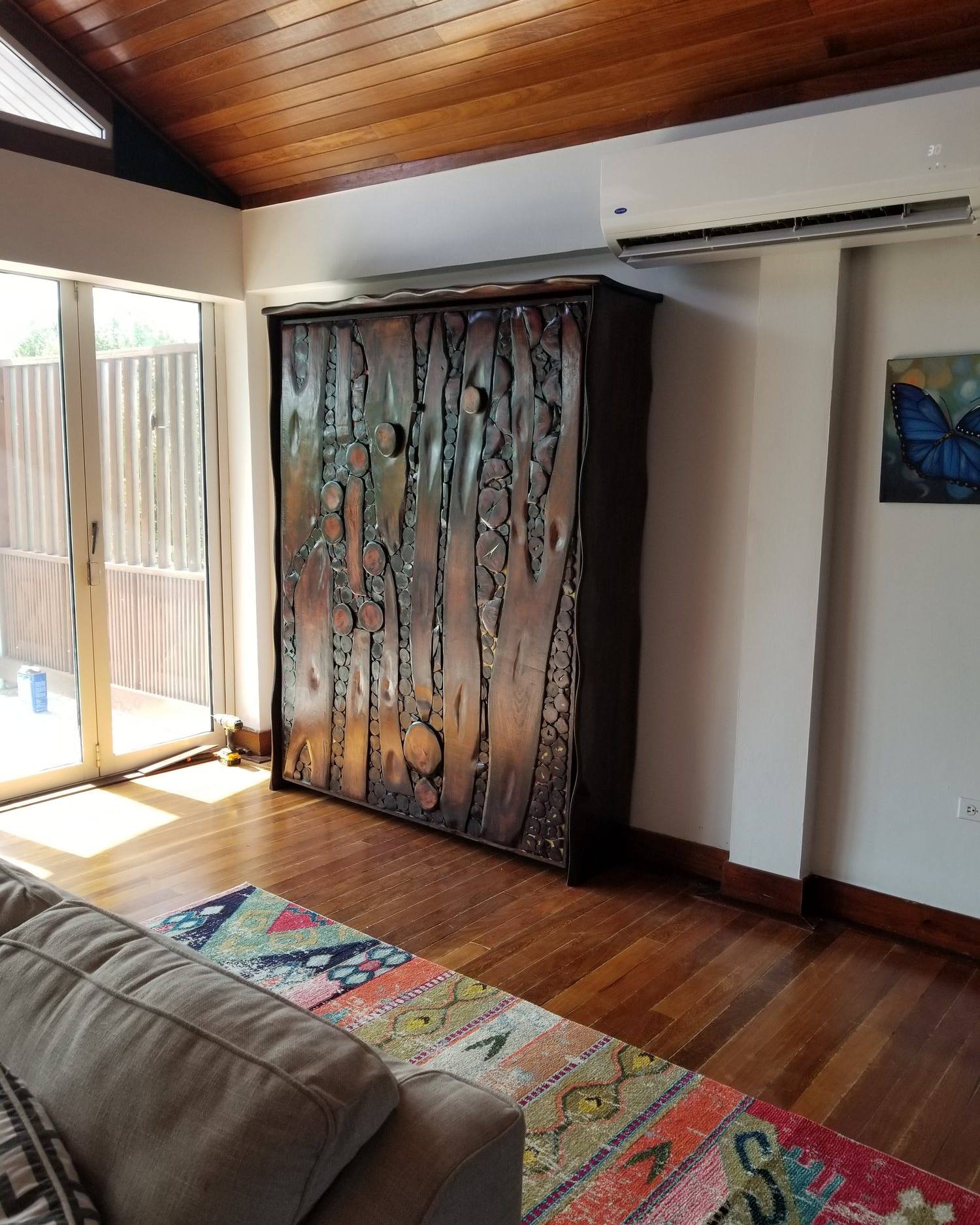 Closed Murphy bed in a living room, its front made of long carved hardwood branch shapes and small rounds fused with resin